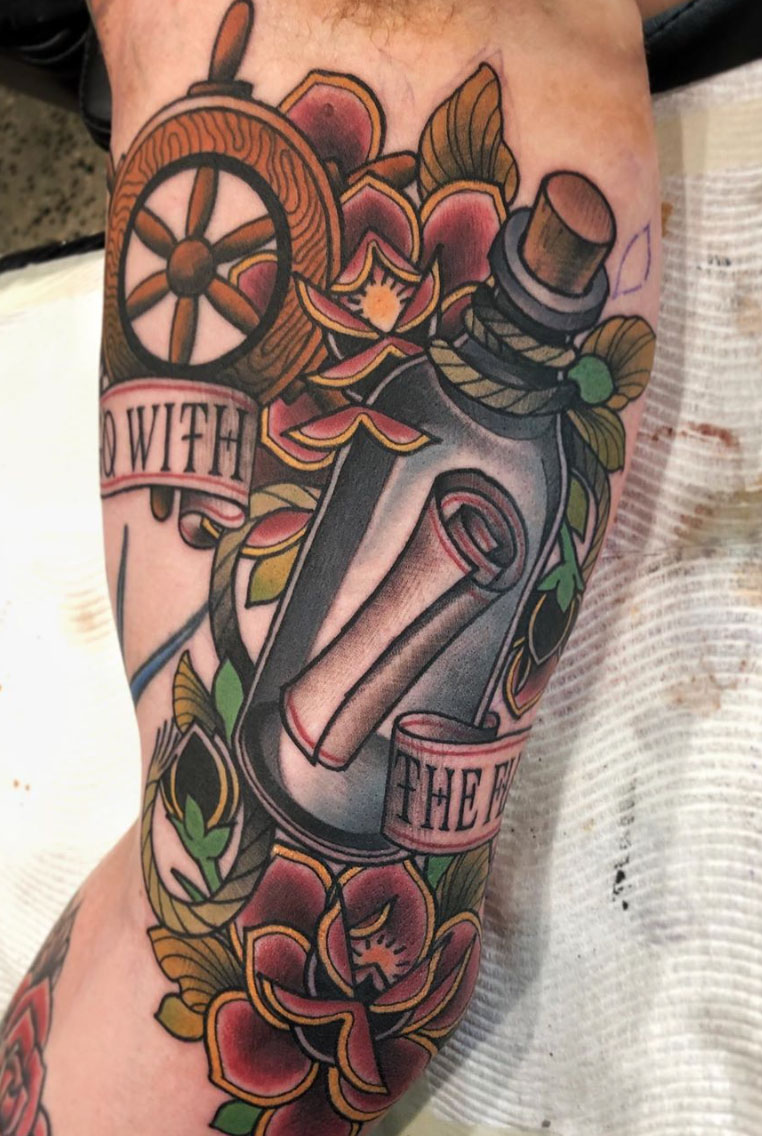 Nautical tattoo with ship's wheel, message bottle, flowers, and banner on person's skin.