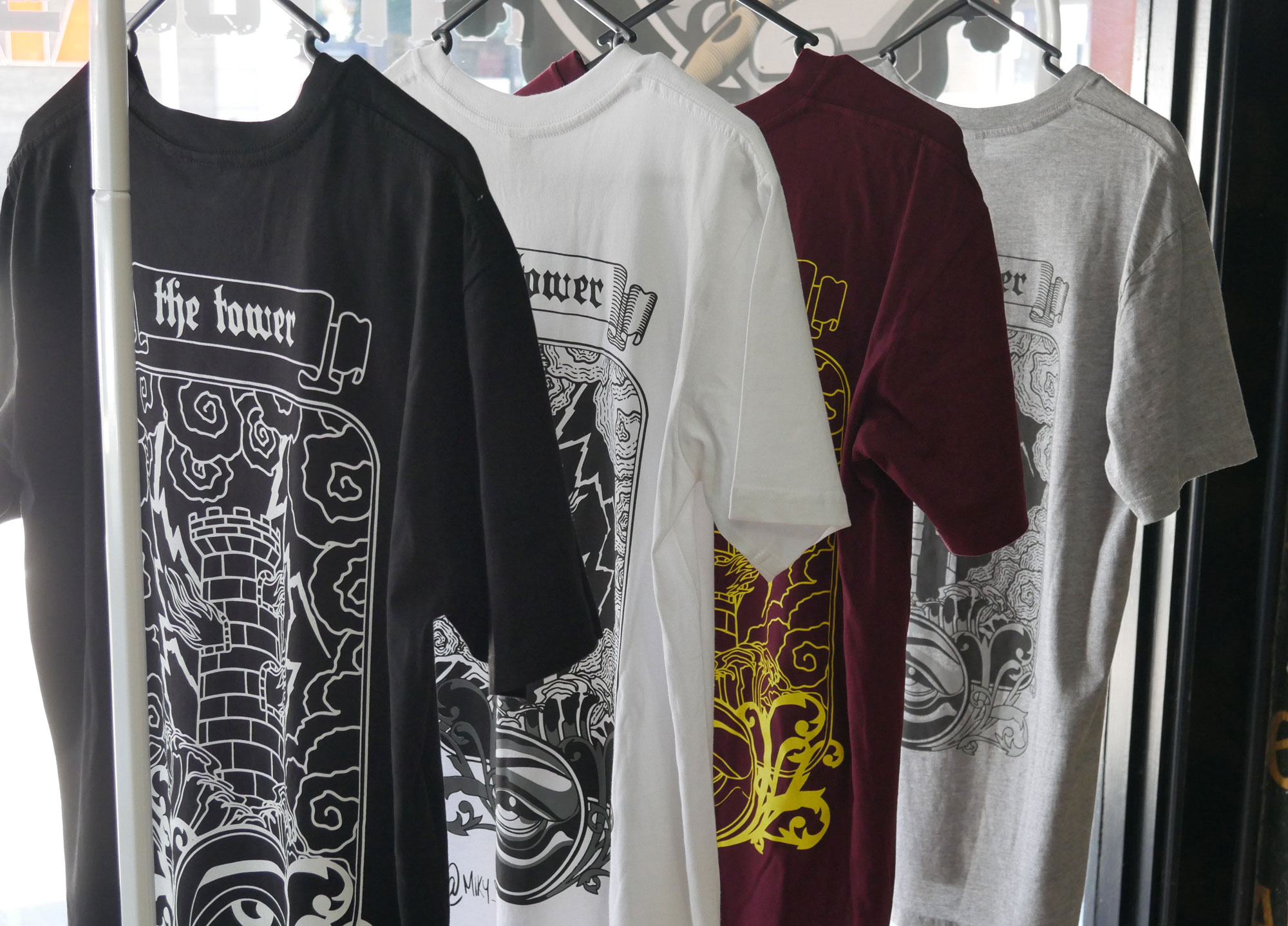 Four t-shirts on hangers with intricate "the tower" prints in black, white, burgundy, grey.