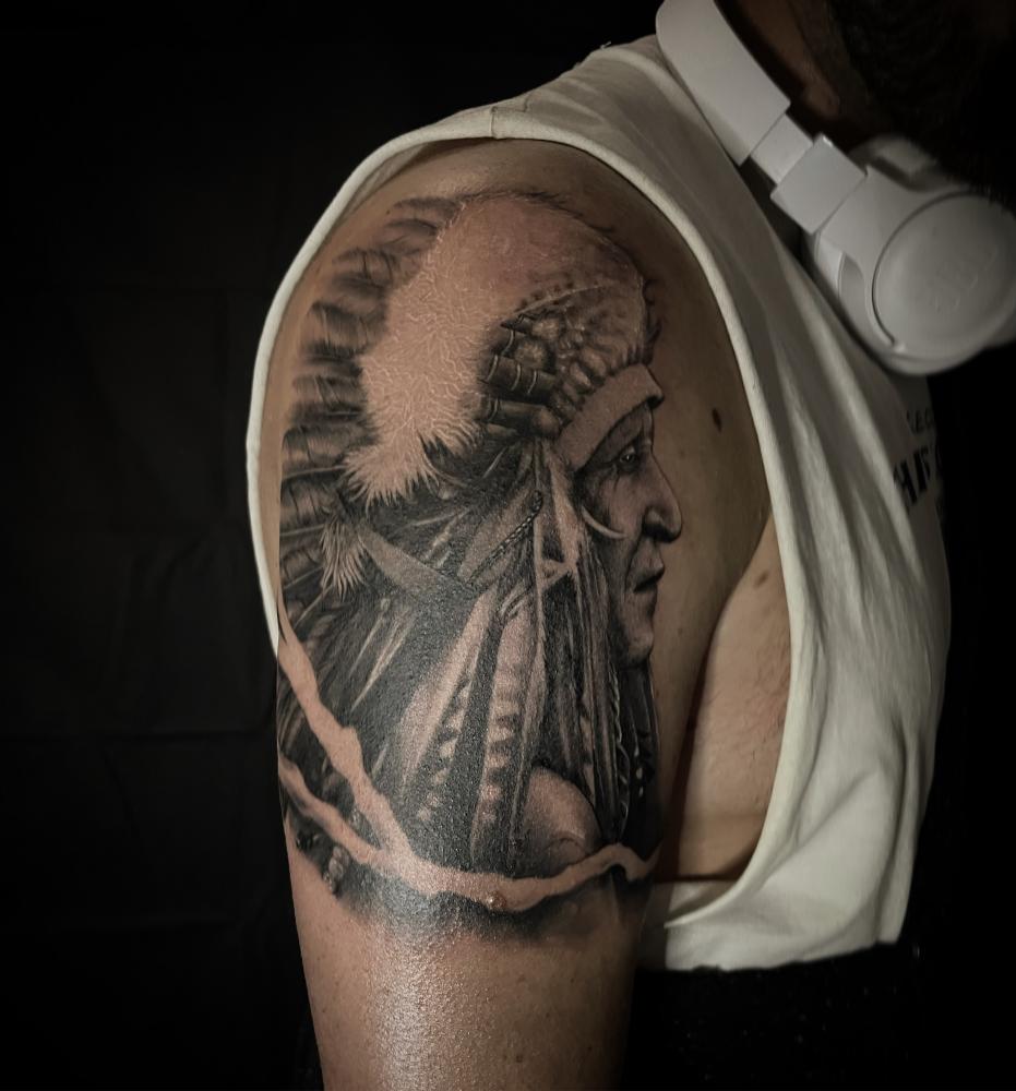 Tattoo of a detailed face with feathered headdress on a person's upper arm.