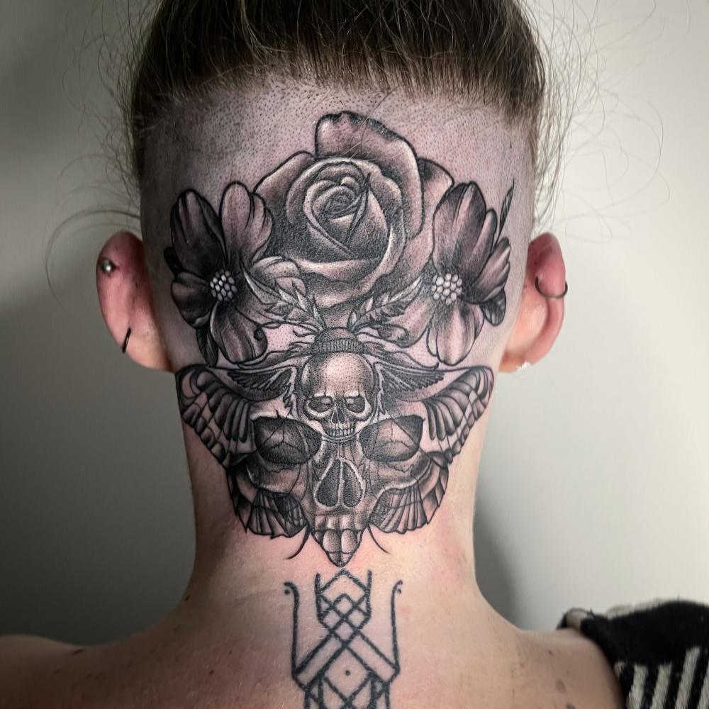 Tattoo of rose, flowers, skull, and moth on shaved head with multiple ear piercings.