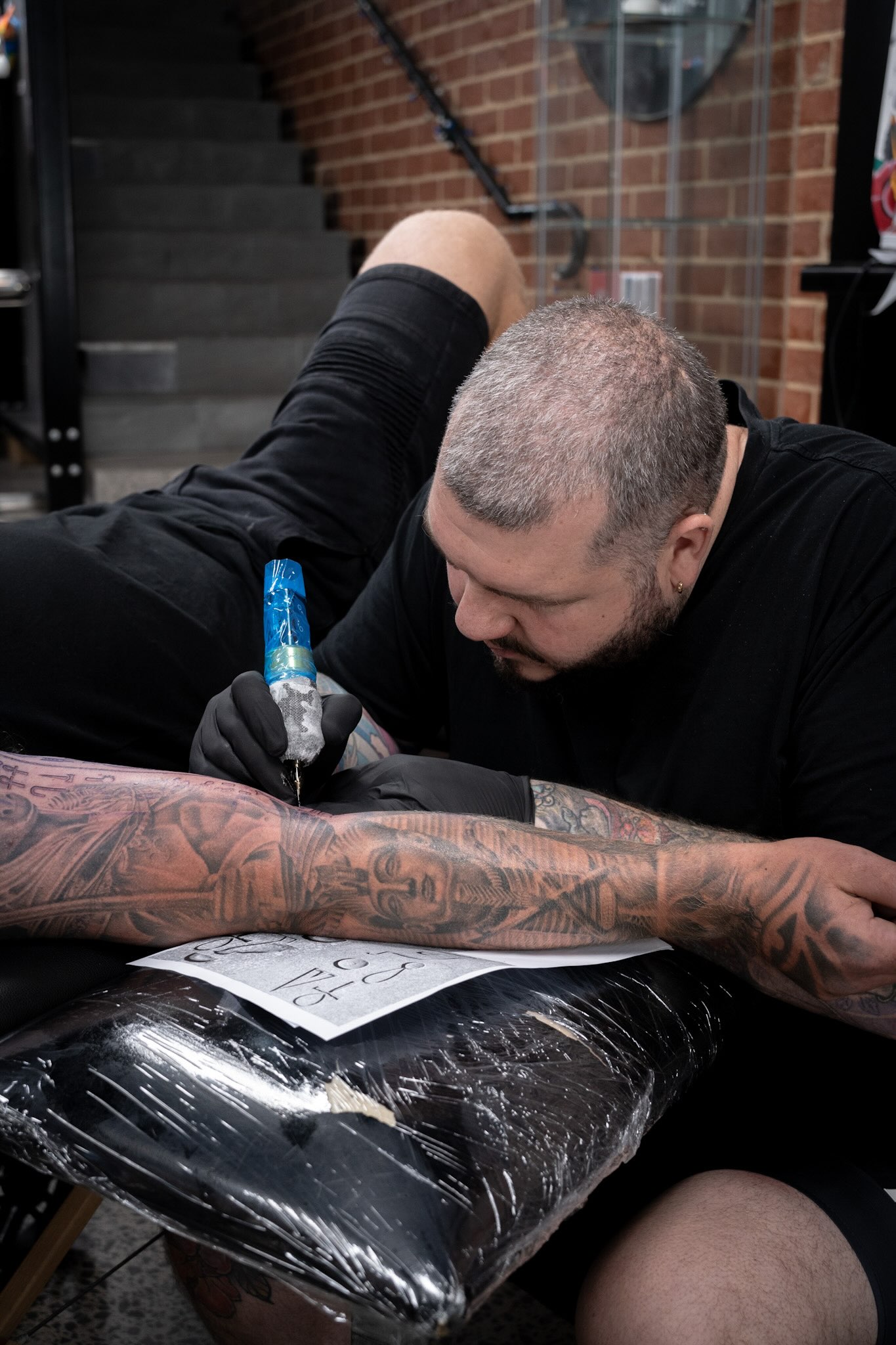 Tattoo artist inking detailed forearm design in studio with sketches, brick wall, staircase.