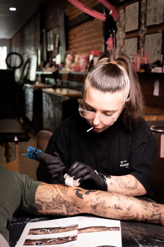 Tattoo artist applying a detailed design to a forearm in a studio setting, wearing gloves.