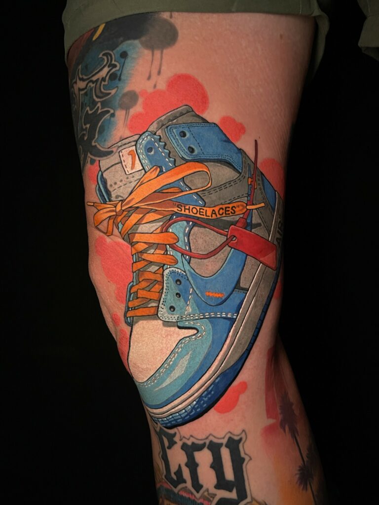 Tattoo of a colorful sneaker with orange laces and "SHOELACES" text on a leg.