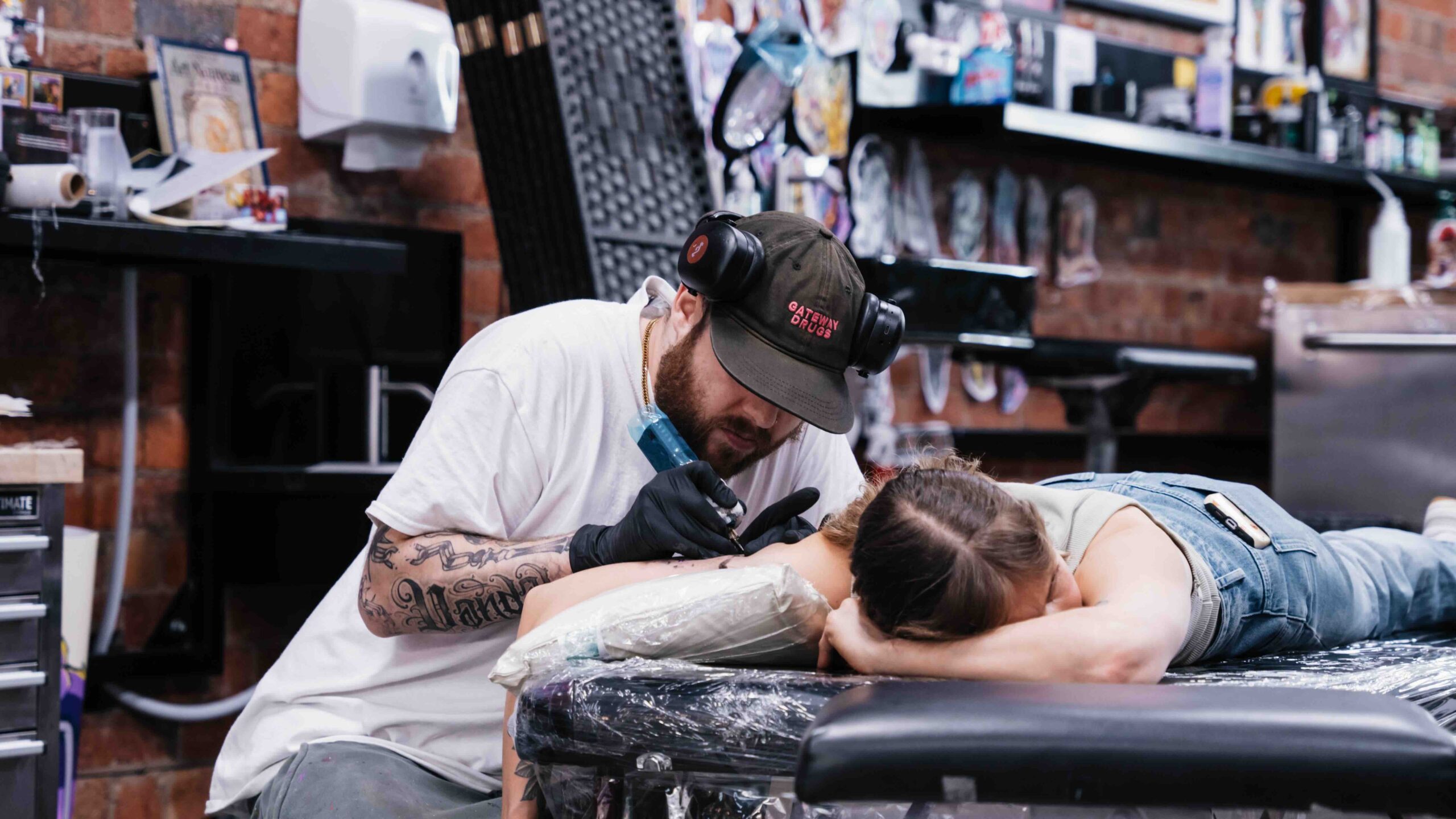 Tattoo artist working on person's back in studio, with supplies and decorations visible.