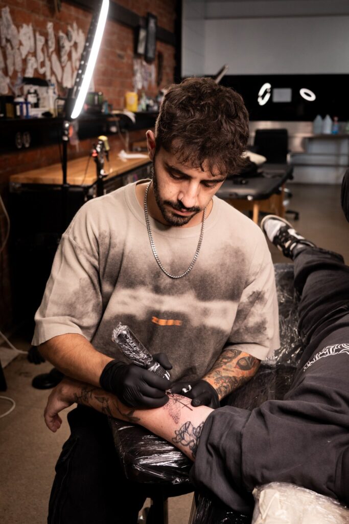Tattoo artist tattooing client's arm in a studio with equipment and artwork on walls.