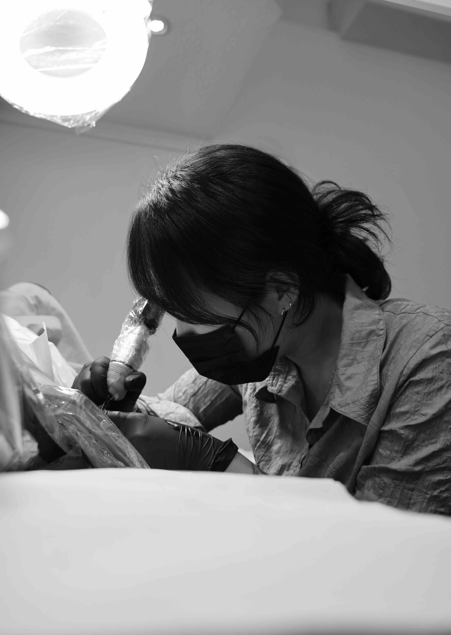 Person in mask and gloves tattooing someone in a well-lit studio with protective covering.
