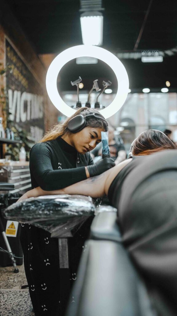 Tattoo artist with headphones works on client's arm under bright studio lighting.