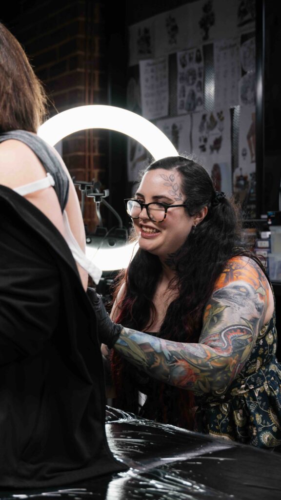 Tattoo artist with facial tattoo smiling while working on a client's back in a well-lit studio.