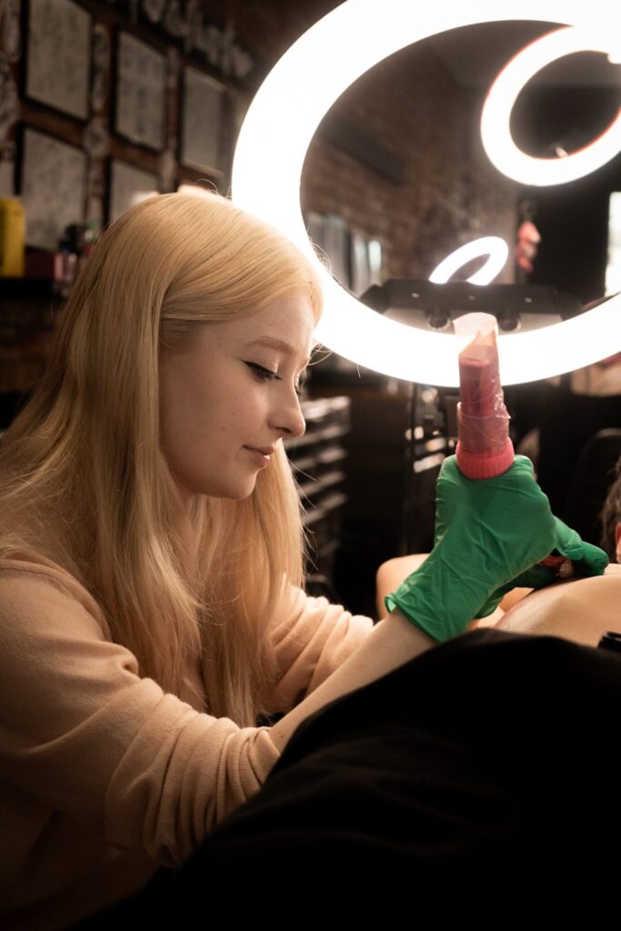 Person with long blonde hair tattooing under a ring light, wearing a green glove.