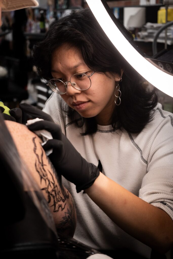 Tattoo artist focused on arm design under bright light, wearing glasses and gloves.