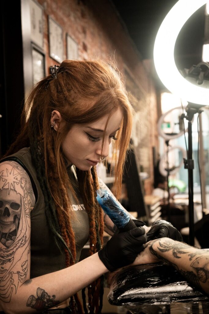 Tattoo artist with auburn hair focused on client's arm, lit by ring light in a studio.