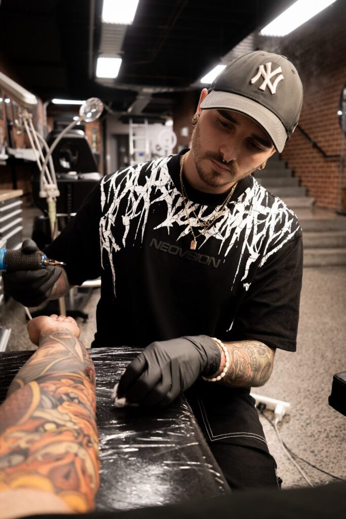 Tattoo artist in cap working on a colorful sleeve tattoo in a modern studio setting.