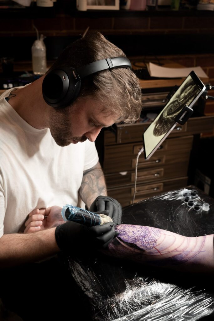 Tattoo artist with headphones tattoos a detailed arm design, using a reference display.