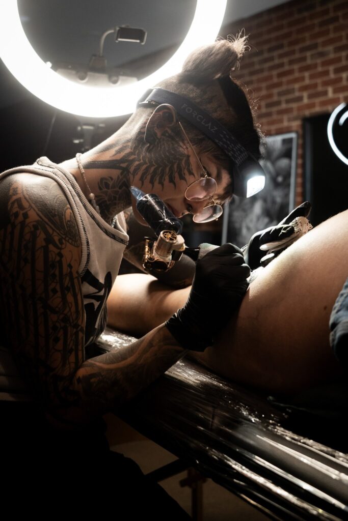 Tattoo artist inking client, wearing glasses and headlamp in well-lit studio.