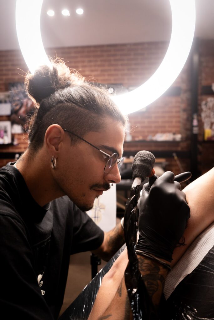 Tattoo artist working on a leg in a well-lit studio with brick wall and equipment.