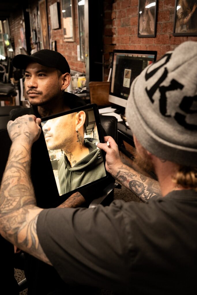 Tattoo artist showing a neck tattoo reference on a tablet to a client in a studio with brick walls.