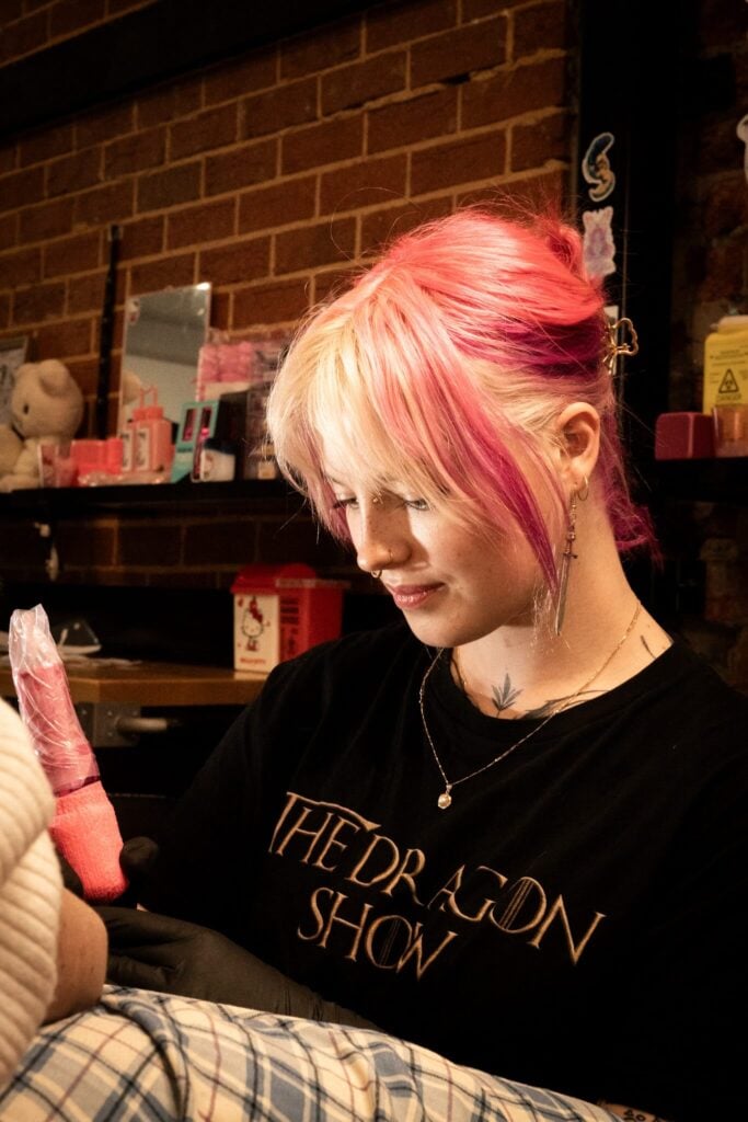 Person with pink and blonde hair tattooing in a room with brick walls and shelves.