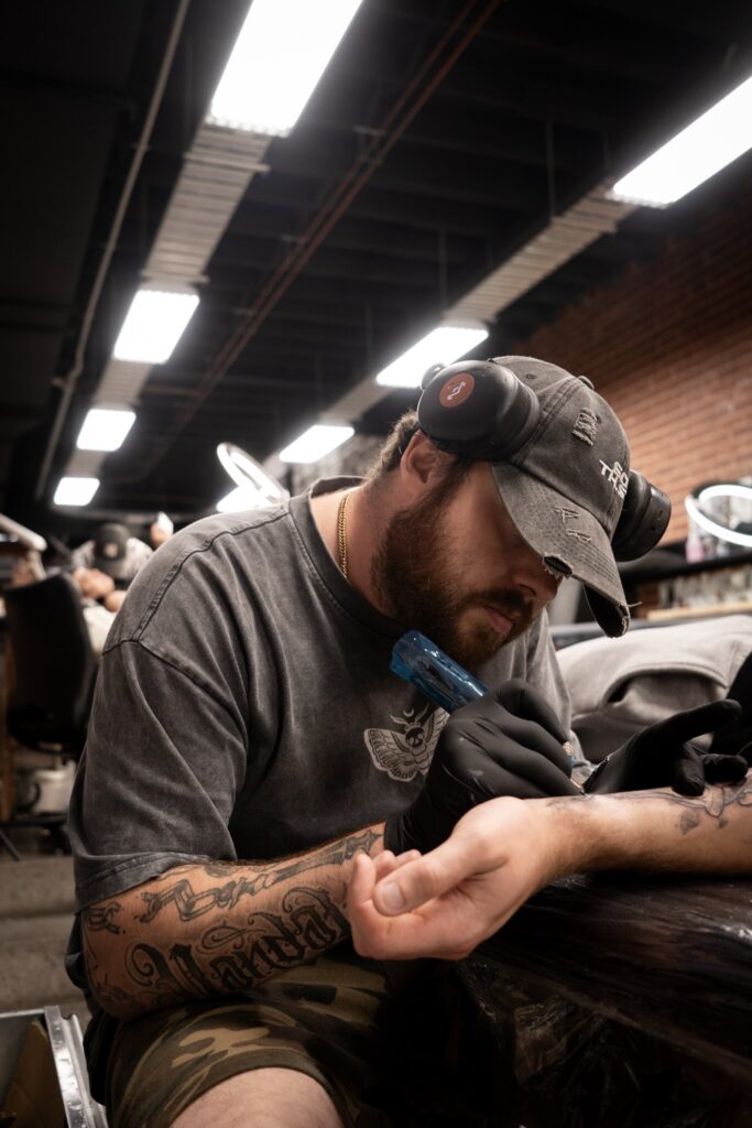 Tattoo artist working on arm in a studio with headphones, cap, and dark clothes, brick wall behind
