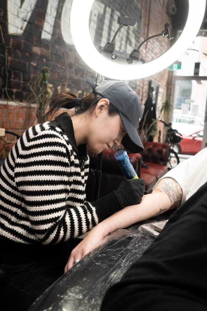 Person in studio tattooing arm under bright ring light with brick wall background.