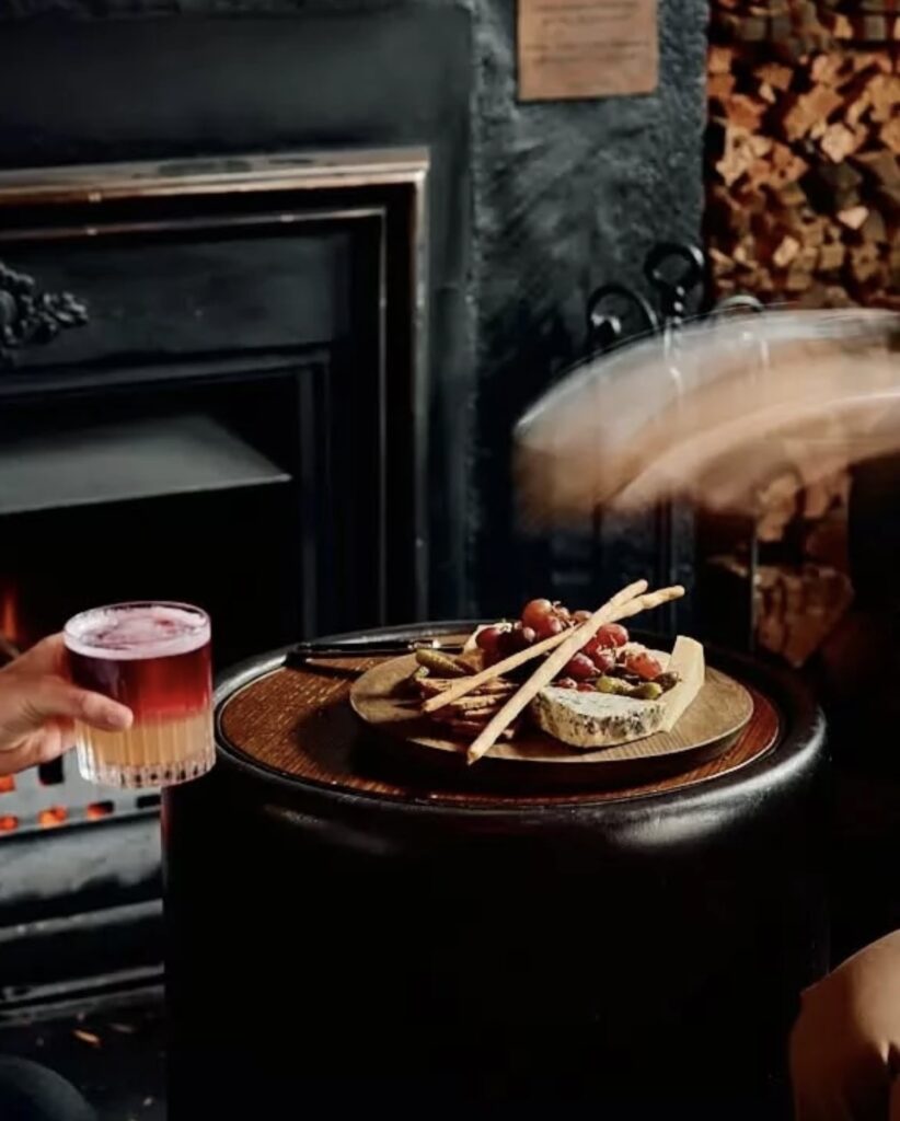 Wooden table with breadsticks, grapes, pickles, and layered drink by a fireplace with firewood.