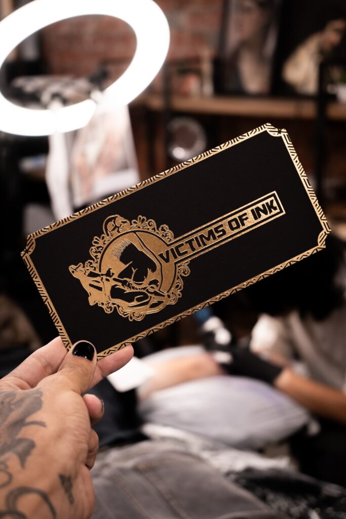 Hand holding "Victims of Ink" card; tattoo studio with person working behind.