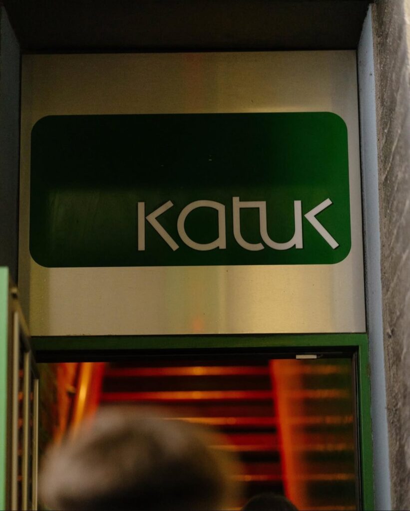 Sign reading "Katuk" on a green background above entrance, red/orange lighting visible below.
