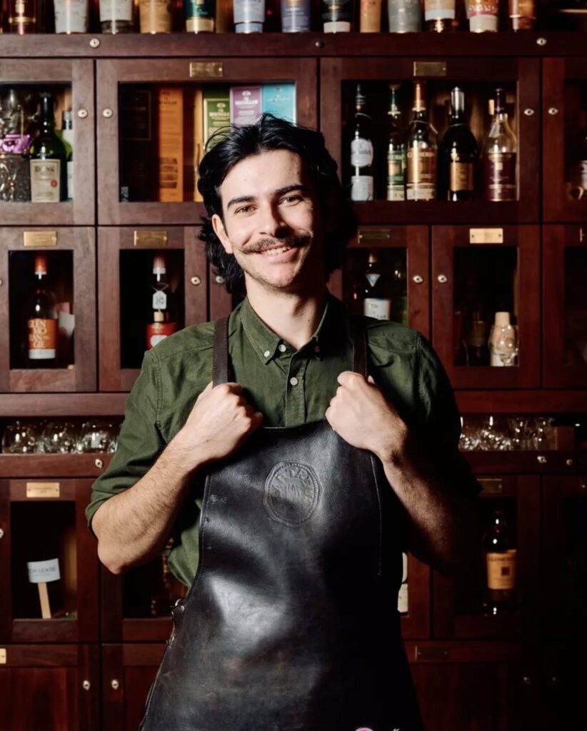 Person in a dark apron and green shirt stands before a wooden cabinet with various bottles.