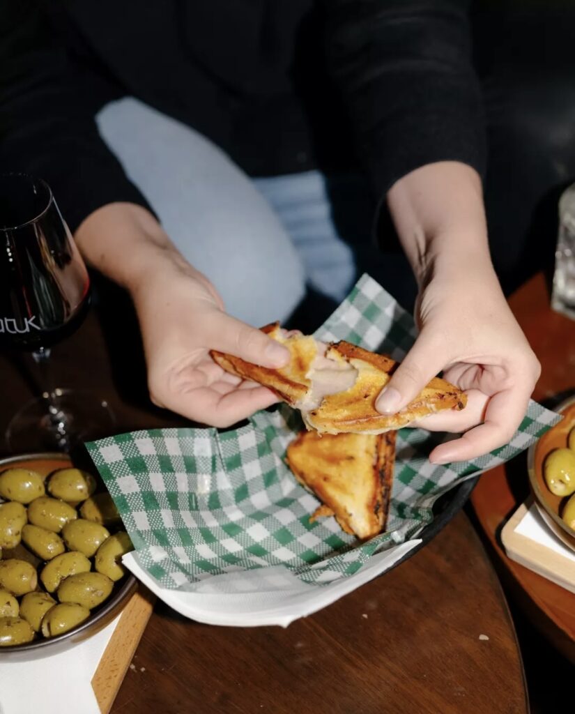Person pulling apart a toasted sandwich over a checkered napkin with olives and wine nearby.