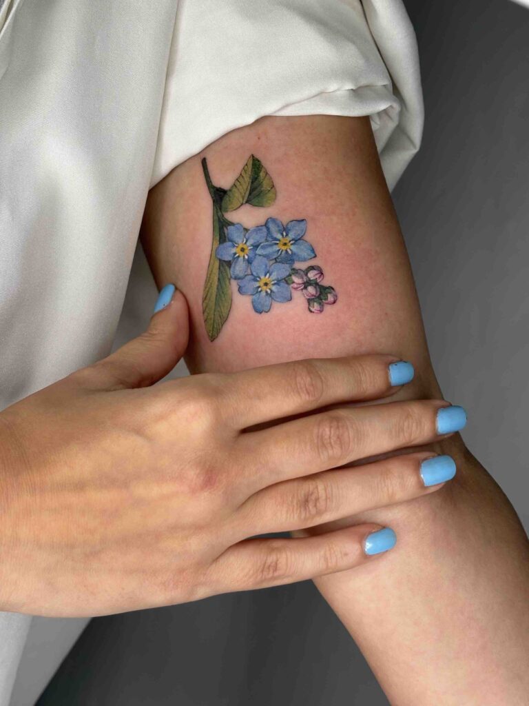 Forearm tattoo of blue flowers and green leaves with a hand showing blue painted nails.