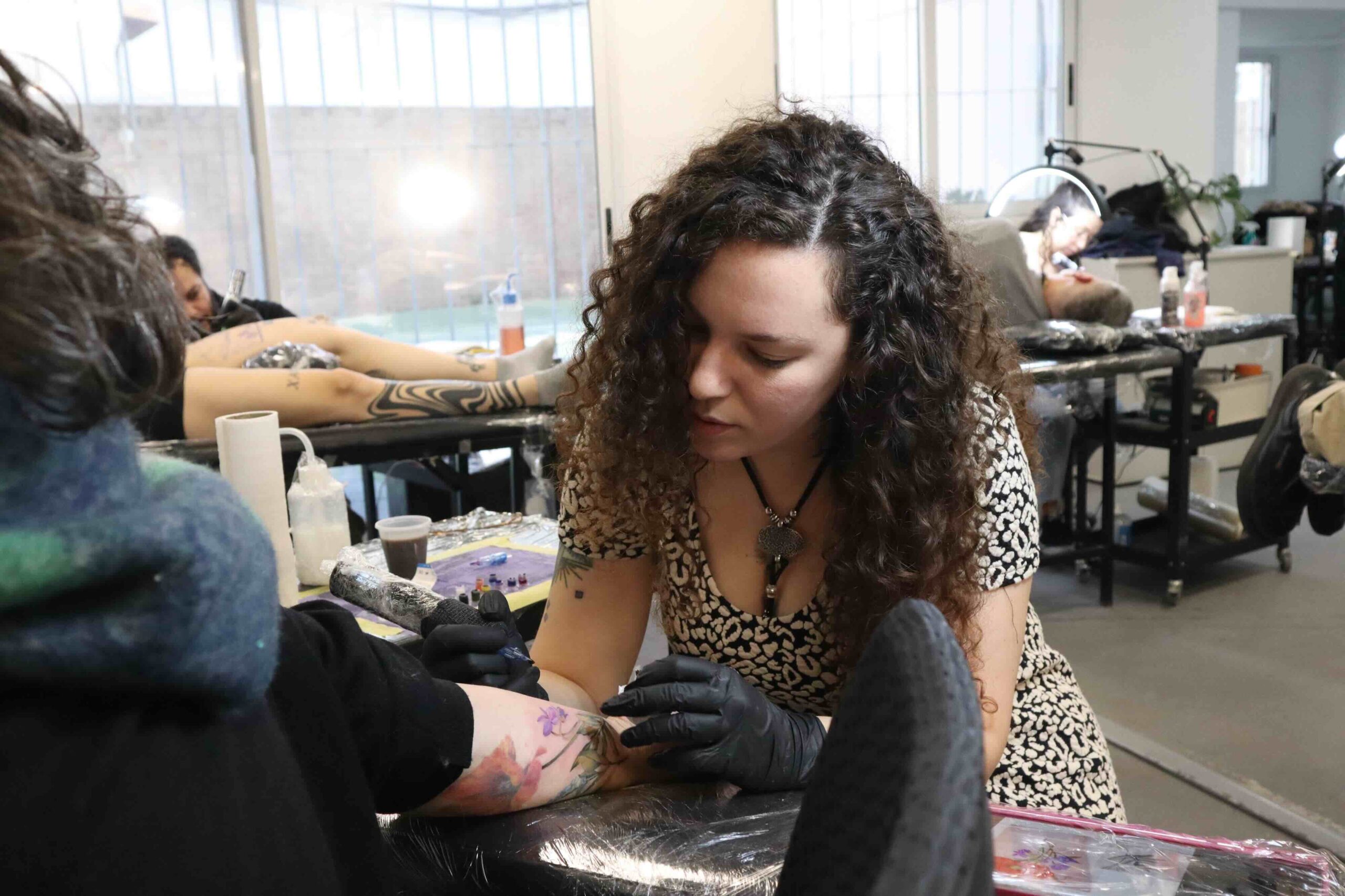 Tattoo artist with curly hair tattooing a floral design on a client's arm in a busy studio.