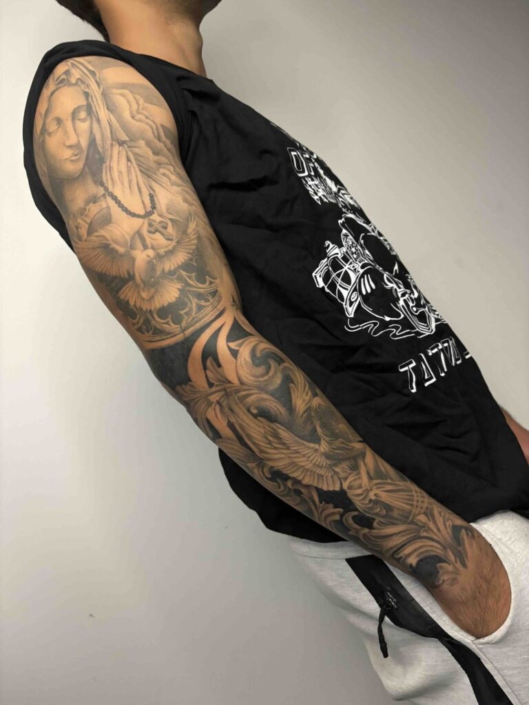 Arm with religious tattoos, dove, rosary; black sleeveless shirt, light pants, hand in pocket.