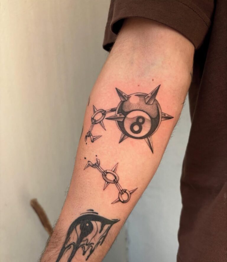 Forearm tattoo of stylized spiked eight-ball and chain-like structure with pointed elements.