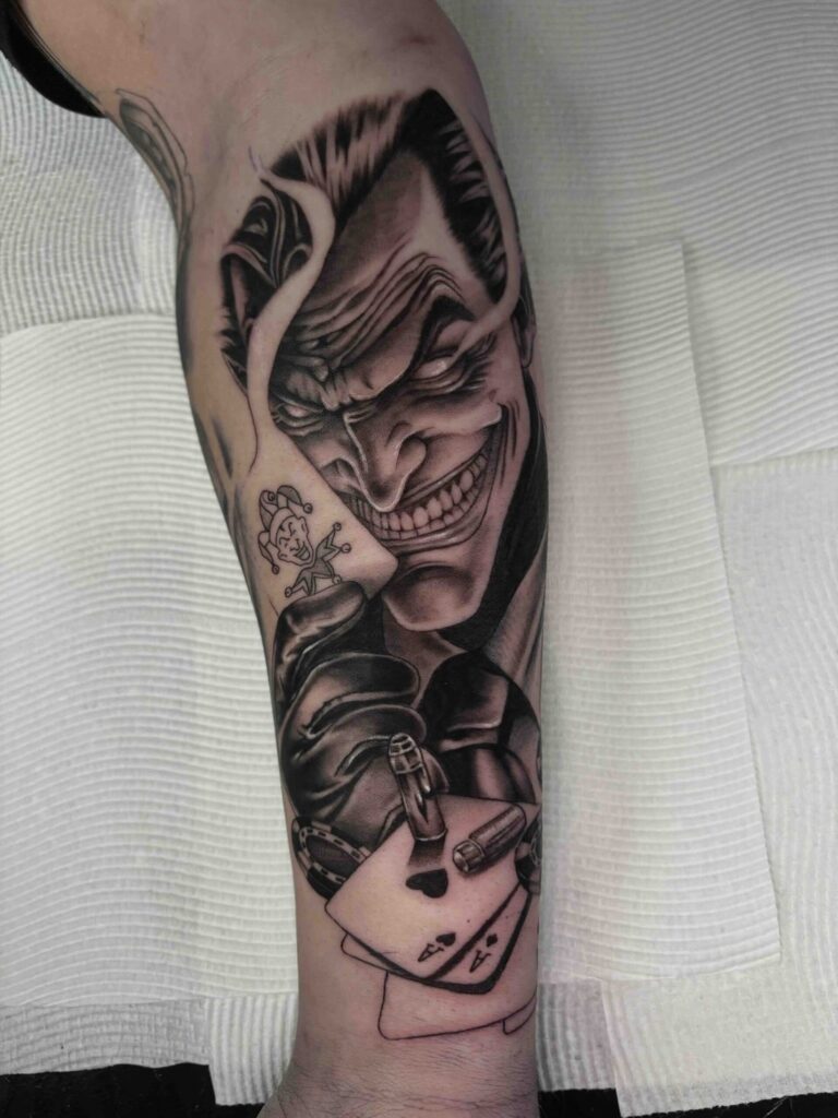 Tattoo on arm features grinning figure, playing cards, and bullets, in black and grey design.