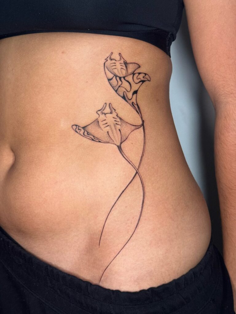 Two fine-line manta rays with flowing tails on the ribs, tattooed by David Carry