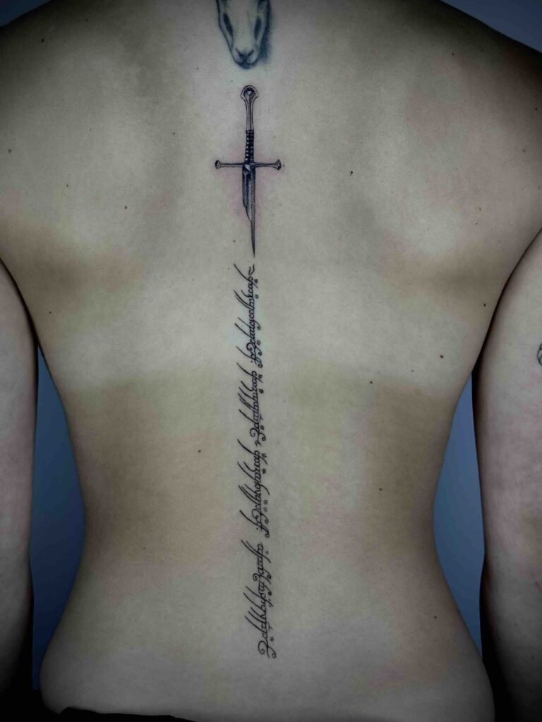Back tattoo featuring an animal head, vertical sword, and decorative script along the spine.