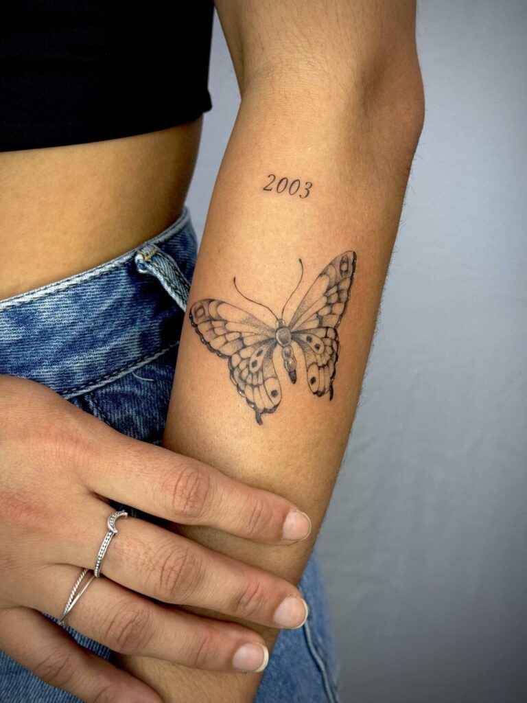 Forearm tattoo of a butterfly with "2003" above, hand in denim jeans and silver ring visible.