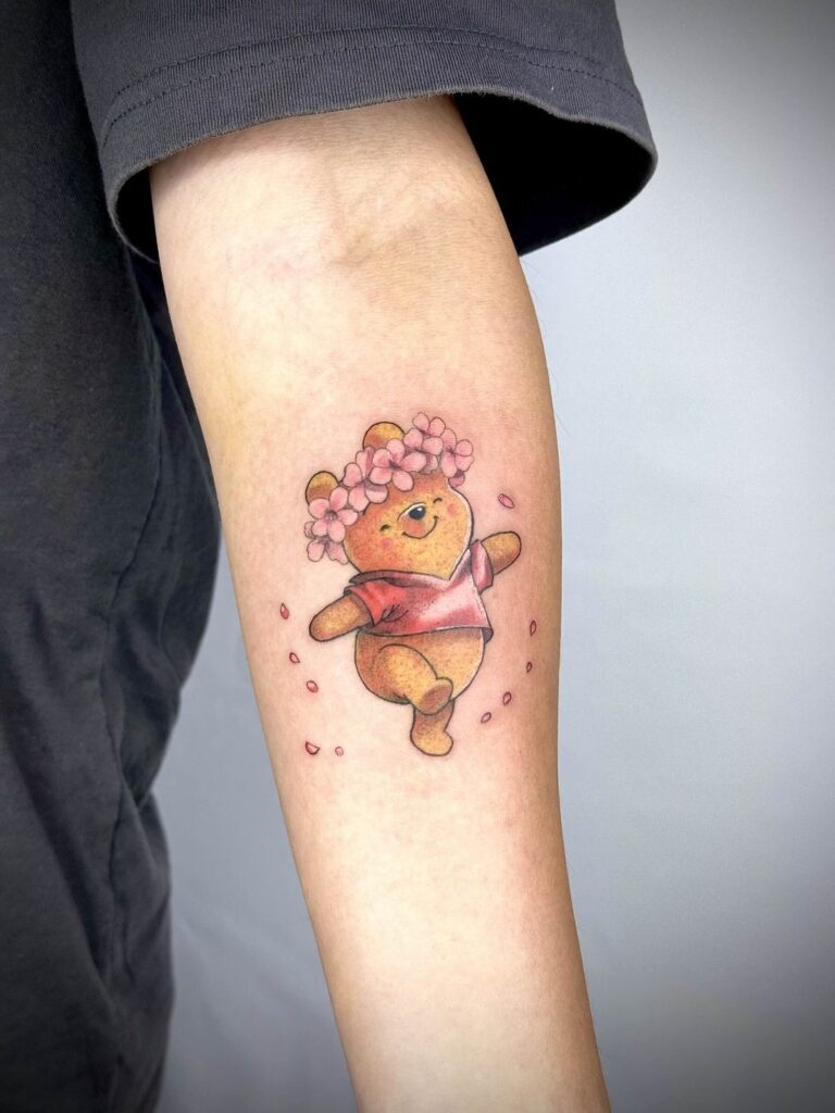 Arm tattoo of a bear in a red shirt, pink flower crown, with surrounding pink petals.