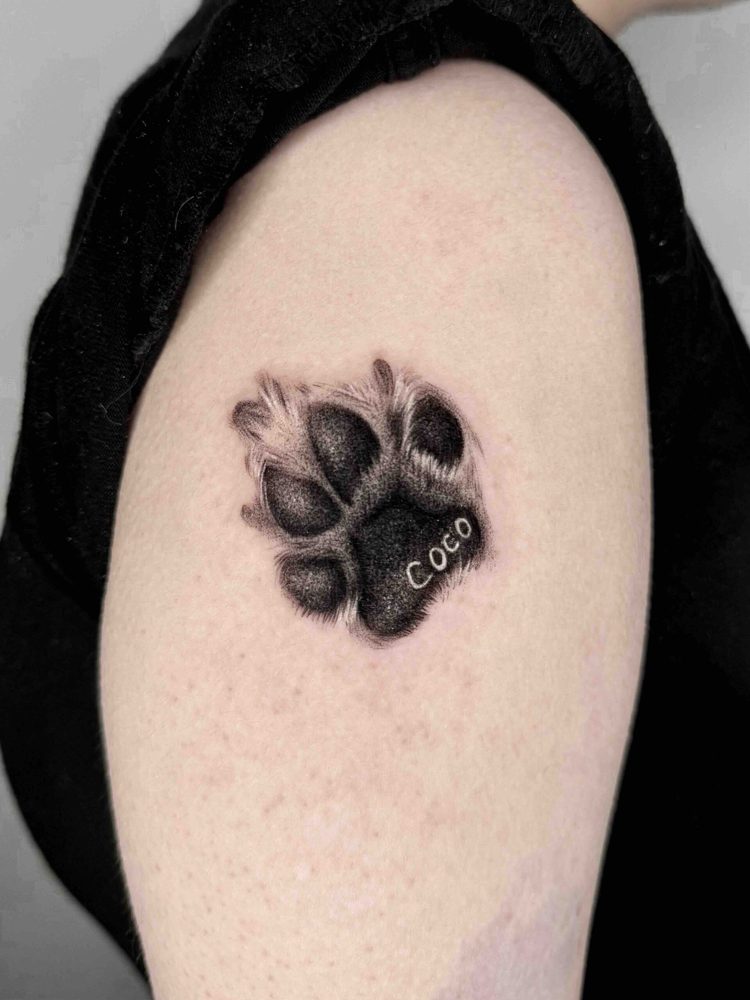 Shaded paw print tattoo with the name Coco on the upper arm by Woody