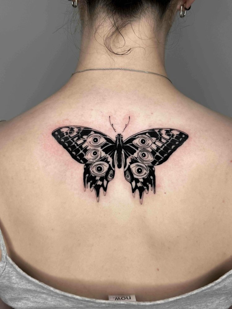 Butterfly tattoo with intricate eye pattern on upper back between shoulder blades.
