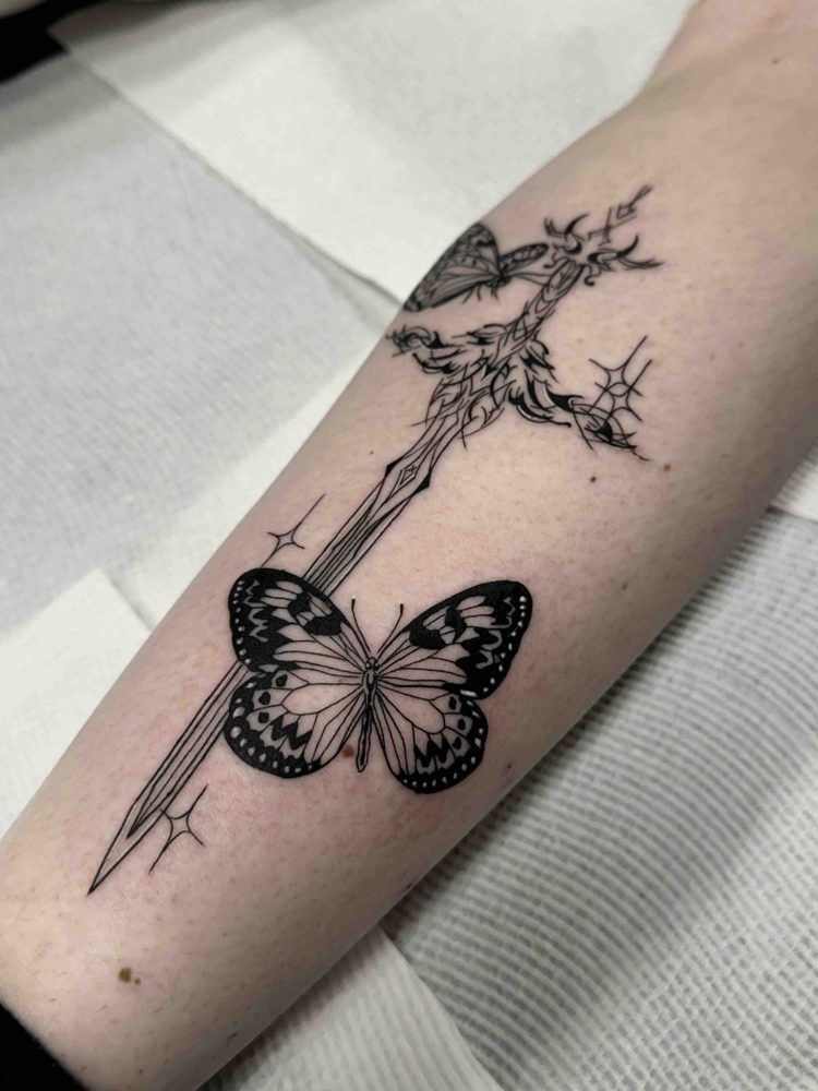 Forearm tattoo of a sword with ornate detailing and two butterflies, featuring intricate patterns.