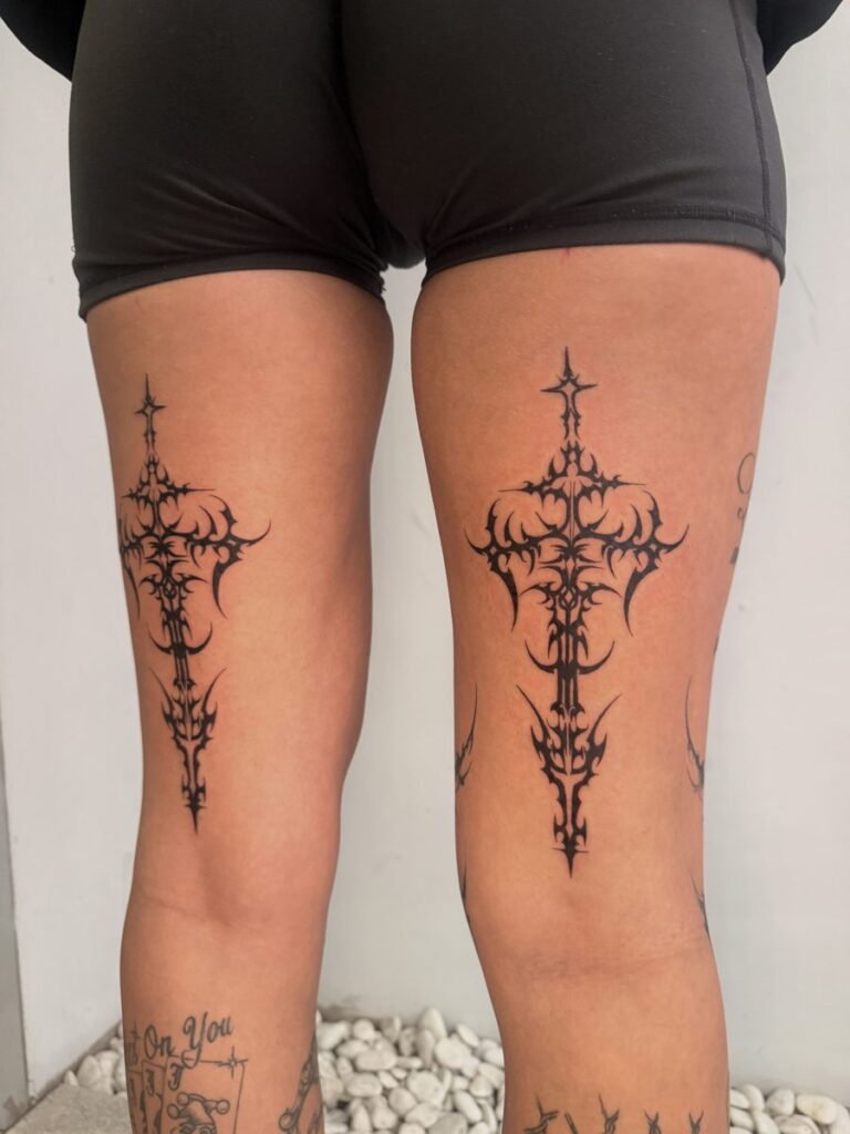 Matching black ornamental cross tattoos on the fronts of both thighs by Chanelle Pititto.