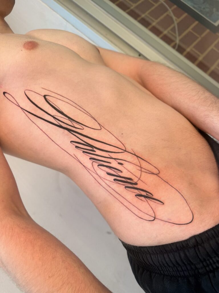Elaborate black script tattoo with long calligraphic flourishes across the ribs by Chanelle Pititto.